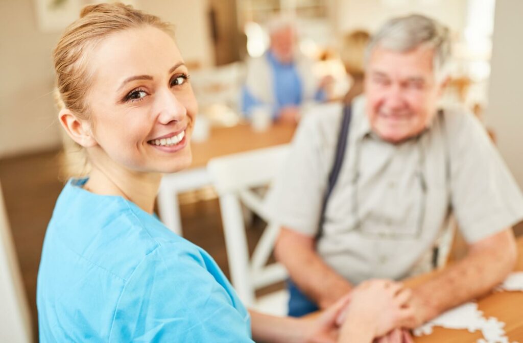 A caregiver checking in on a resident turns to smile at the camera while kneeling in the lounge of an assisted living community