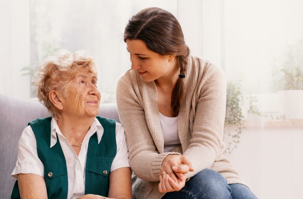 A caregiver sits on the arm of a couch and smiles down at a happy seated resident in senior living