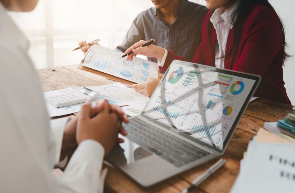 3 financial advisors sit together during a meeting to review printed reports. A laptop holds data, graphs, and charts with financial information.