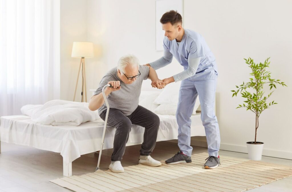 A caregiver helping a senior man with a cane stand up from a bed.