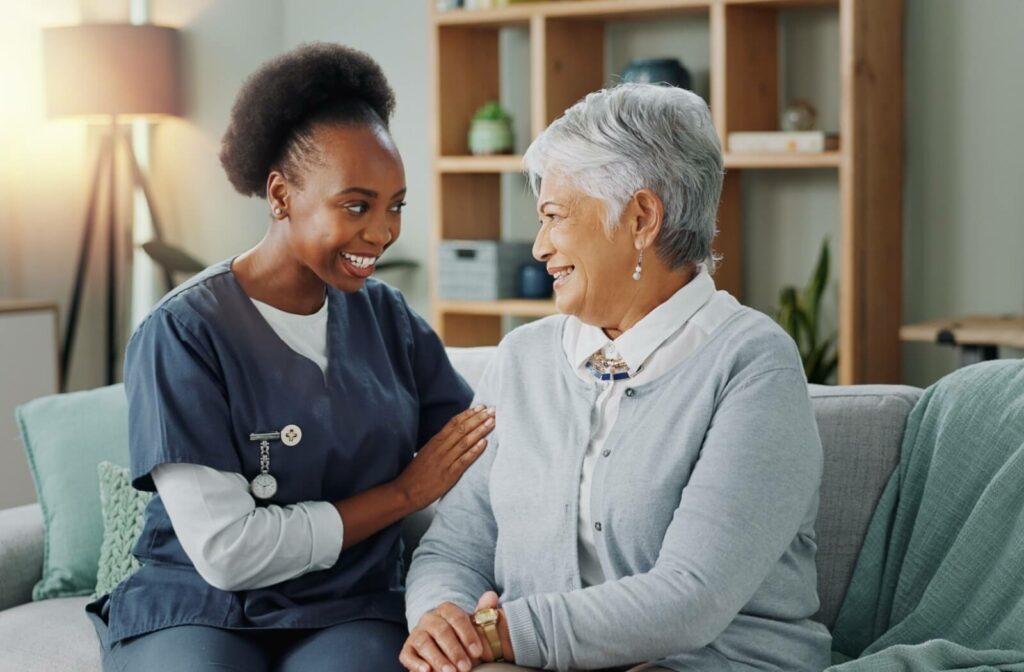 A caregiver and resident in senior living sit on a couch and smile at one another.