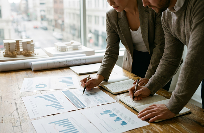 Two executives, in a conference room, analyze data charts on a wooden table with an architectural model of a senior living community in the background.